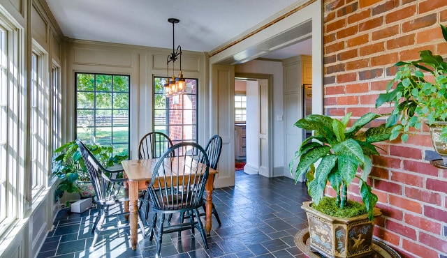 Sunroom with one brick wall and three white-painted walls with large windows, featuring a wooden table and chairs and a variety of plants.