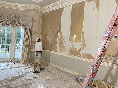 Progress photo of a living room remodel featuring a man stripping wallpaper from the wall.