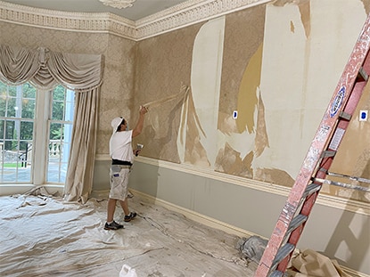 Progress photo of a living room remodel featuring a man stripping wallpaper from the wall.