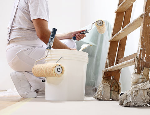 A painter kneeling next to a bucket and ladder surveys his work, paint roller in hand. A painter kneeling next to a bucket and ladder surveys his work, paint roller in hand.