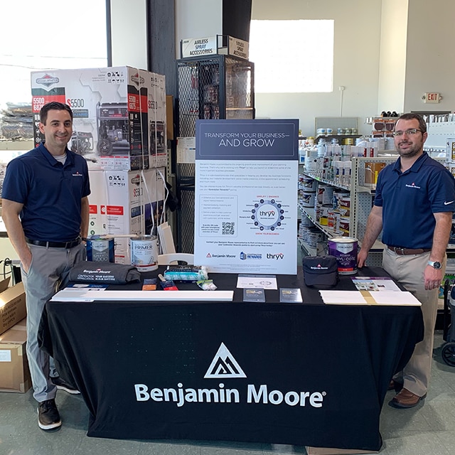 Deux hommes portant des polos Benjamin Moore se tiennent de chaque côté d’un présentoir d’éléments promotionnels et d’un panneau Thryv dans un magasin Benjamin Moore indépendant.