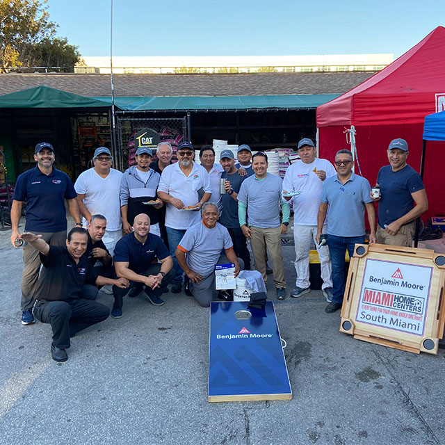 A group of men posing outside near red and blue tents with Benjamin Moore merchandise displayed on a table and a blue cornhole board.