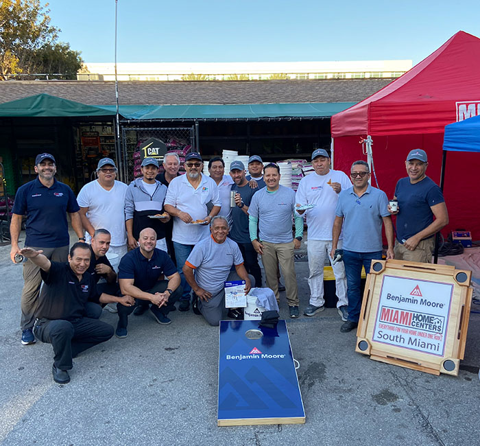 A group of men posing outside near red and blue tents with Benjamin Moore merchandise displayed on a table and a blue cornhole board.