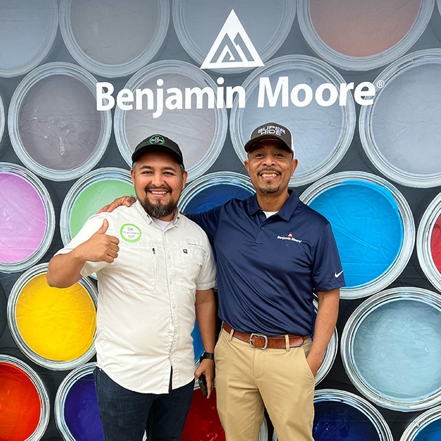 Two men posing in front of a Benjamin Moore backdrop with colorful open cans of paint.