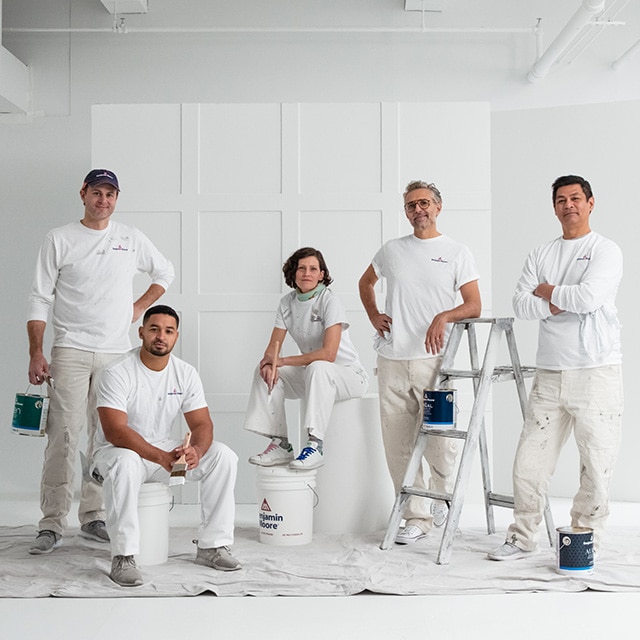A group of five Benjamin Moore painting contractors dressed in white, posing with gallons of paint and a ladder in a white-painted room.