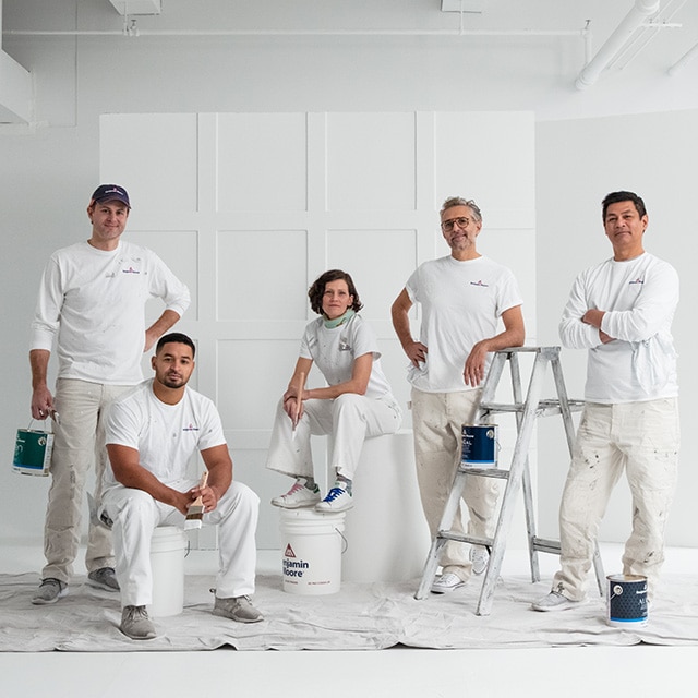 Un grupo de cinco contratistas de pintura de Benjamin Moore vestidos de blanco, posando con galones de pintura y una escalera en una habitación pintada de blanco.