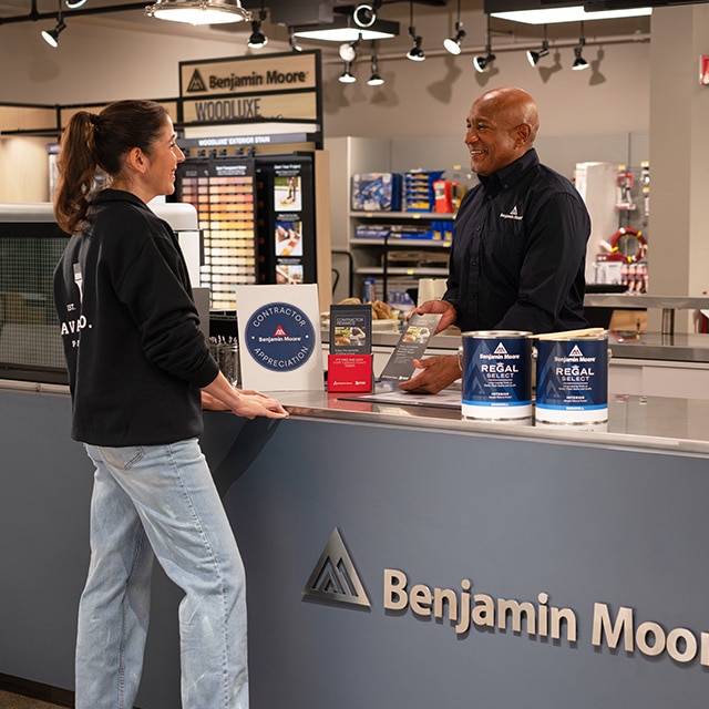 A man and woman in a locally owned Benjamin Moore store standing at a counter with Regal® Select gallons of paint and Contractor Rewards brochures.