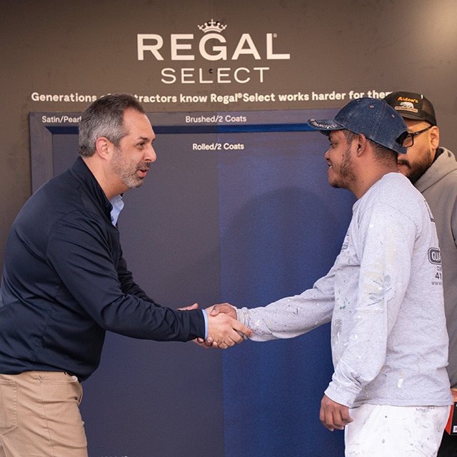 Two men shaking hands in front of a Benjamin Moore Regal® Select backdrop with cans of paint and a panel painted two shades of blue.