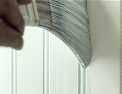 Using a paint brush, a professional painter brushes ben Interior paint in light green to a grooved wall.