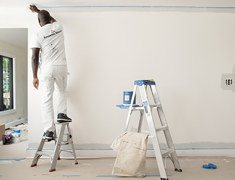 A professional painter uses a step ladder, with a taller ladder on reserve that holds a can of REGAL® Select Interior paint.