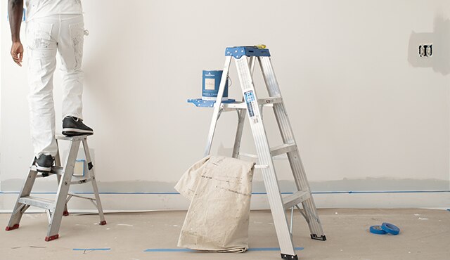 Un peintre professionnel est grimpé sur un escabeau et il en utilise un autre, plus grand, pour y déposer son contenant de peinture d’intérieur REGALMD Select.