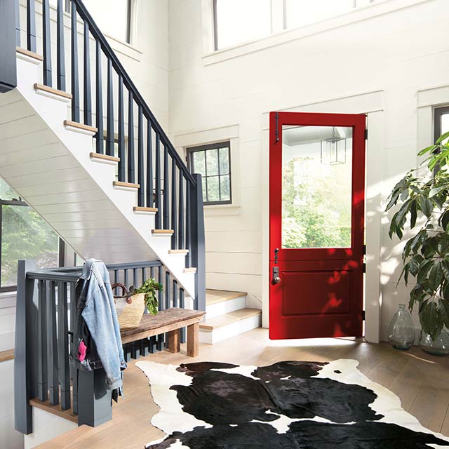 The interior of a home with a deep red-painted front door opening into the white-painted entryway with staircase.