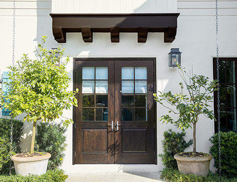 A stucco home with wooden doors and trellis. Bushes and trees adorn the lawn. 