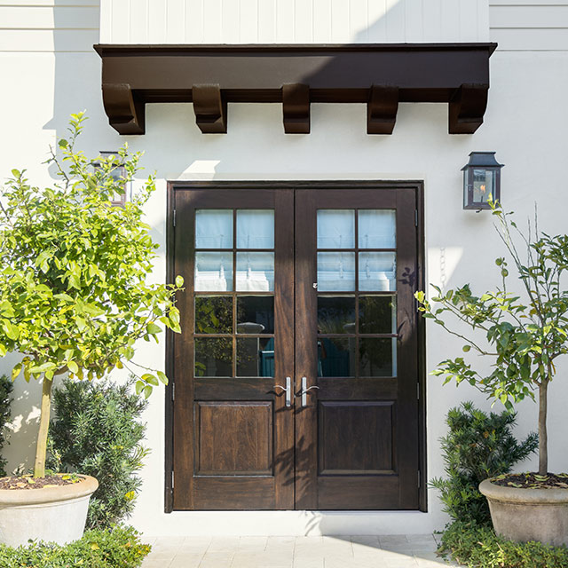 A stucco home with wooden doors and trellis. Bushes and trees adorn the lawn. 