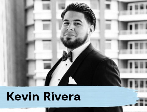 A man with slicked back hair and a full beard wearing a tuxedo with a bow-tie poses in front of a tall, residential building.