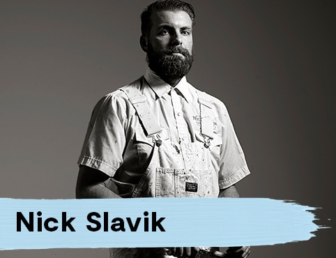 A dark-haired man with a full beard stands in a white, short-sleeved collared shirt, denim overalls, looking ahead.