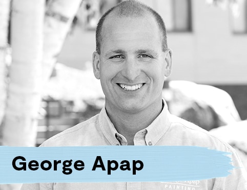 Man with closely cropped hair standing outside an office building, with a button-up shirt labeled “George Apap Painting.”
