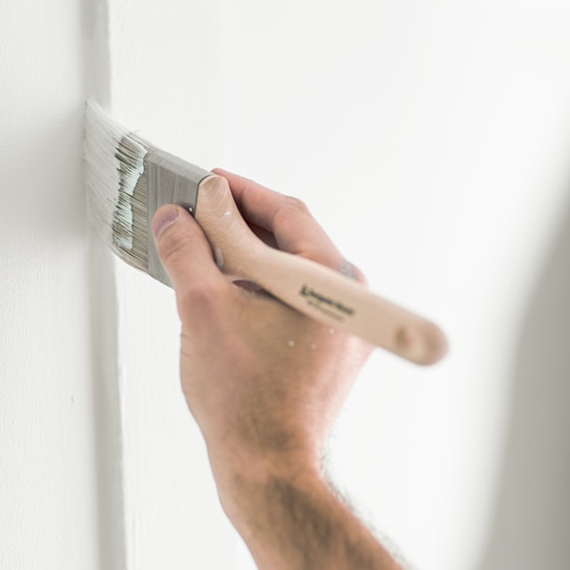 A hand holding a paintbrush paints a wall with white paint.