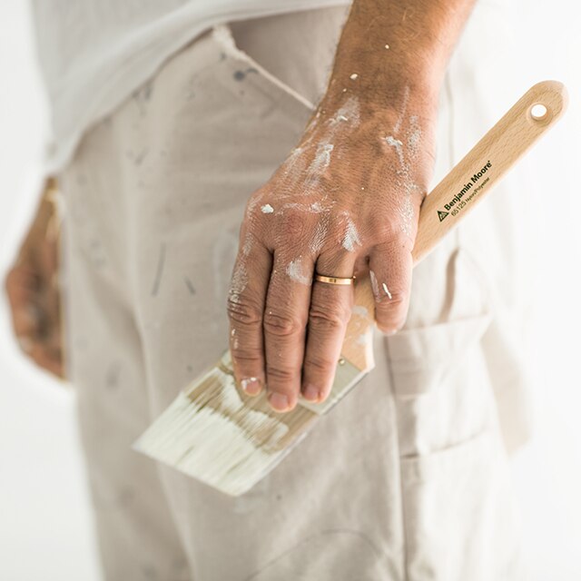 A side shot of the lower half of a painting contractor shows a paint-covered left hand holding a paintbrush.