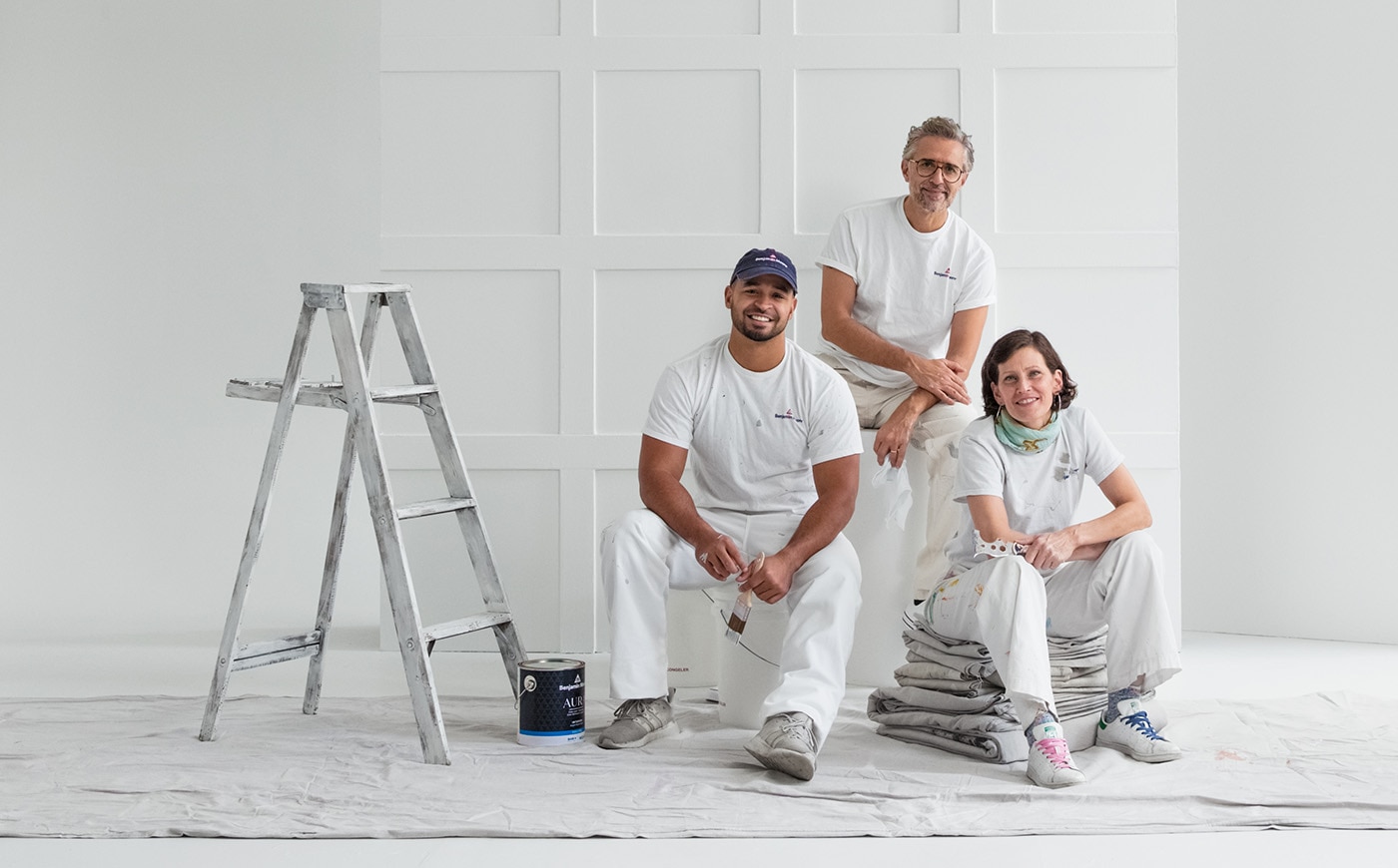Two male and one female painting contractors sit on a drop cloth on the floor of an industrial-looking, all-white room with black window trim.