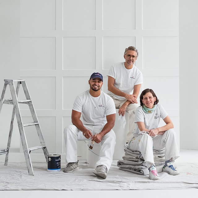 Two male and one female painting contractors sit on a drop cloth on the floor of an industrial-looking, all-white room with black window trim.