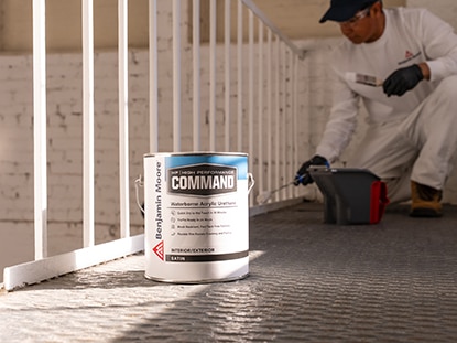 A gallon of Benjamin Moore Command® Waterborne Acrylic Urethane next to a painting contractor painting a white railing.
