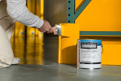 A person kneeling and brushing orange paint onto a section of wall behind a gallon of Benjamin Moore Command® Waterborne Acrylic Urethane. 