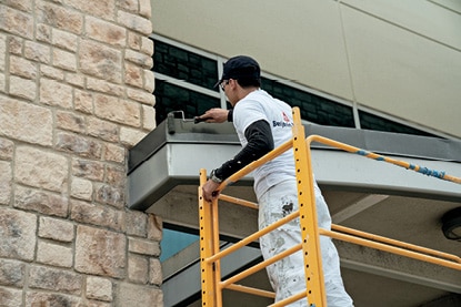 A Benjamin Moore painting contractor standing on yellow scaffolding and painting a facility exterior with Corotech® COMMAND®.