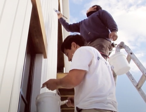 Two professional contractors painting the exterior of a house with white paint, with one bucket of paint hanging on a ladder.