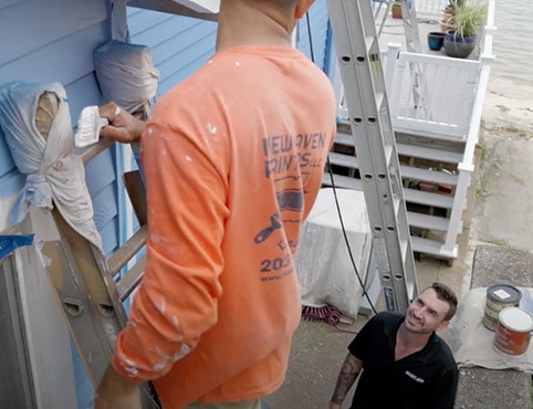 A painter on a ladder wearing an orange shirt, painting blue siding on a house, with the painting contractor.