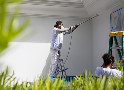 A painter standing on a ladder spray painting the exterior of a house with white paint, a second painter holding a ladder.