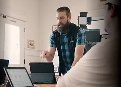 A person pointing at a computer tablet, a second person sitting in front of a tablet.