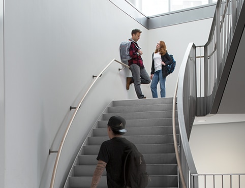 Un homme et une femme se parlent sur le palier d’un escalier sur plusieurs étages avec des murs peints en gris et de grandes fenêtres.