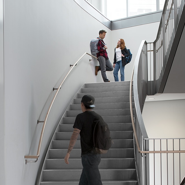 Un homme et une femme se parlent sur le palier d’un escalier sur plusieurs étages avec des murs peints en gris et de grandes fenêtres.