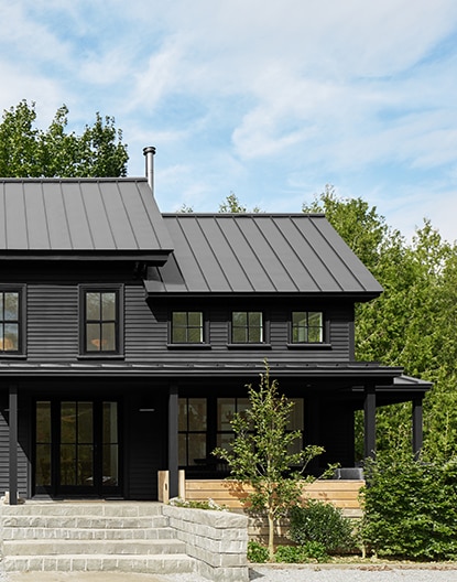 Gray stone masonry leads up to a modern home with black siding and shed-style aluminum black roofing.
