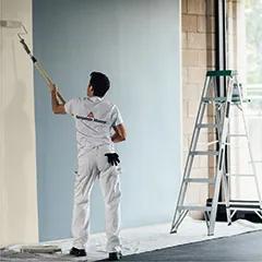 Miniatura: Un contratista de pintura profesional aplica pintura blanca con un rodillo sobre una pared interior azul.