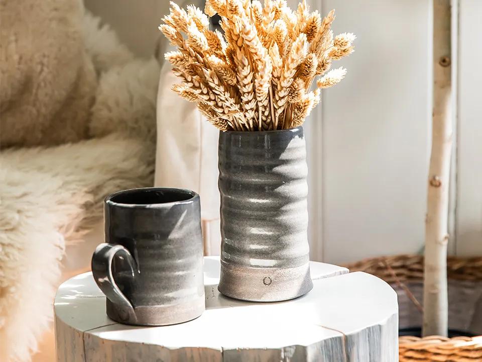 Un jarrón con plantas disecadas y una taza de cerámica marrón sobre una mesa de madera.