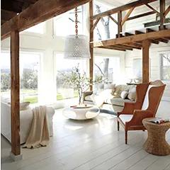 Miniatura: Una sala de estar luminosa pintada de blanco con ventanas altas, vigas de madera natural, un área de loft y decoración informal.
