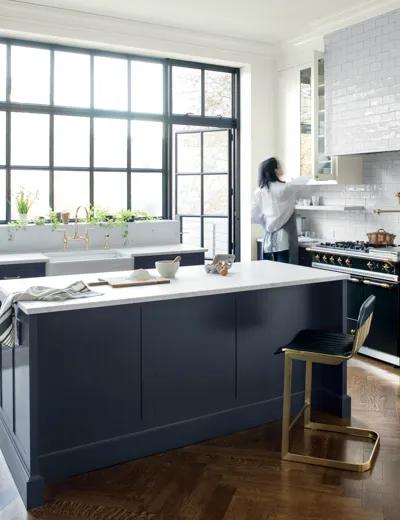 Una cocina blanca con gabinetes inferiores e isla central pintados en azul marino con encimeras blancas, un gabinete de vidrio superior blanco y ventanas grandes.