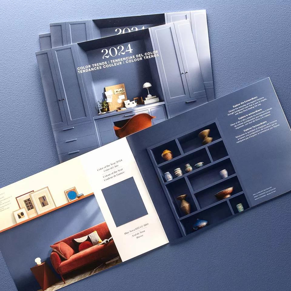 Una pila de folletos del color del año 2024 de Benjamin Moore, con el de arriba abierto en una página central que muestra una muestra de pintura Azul de Nova.