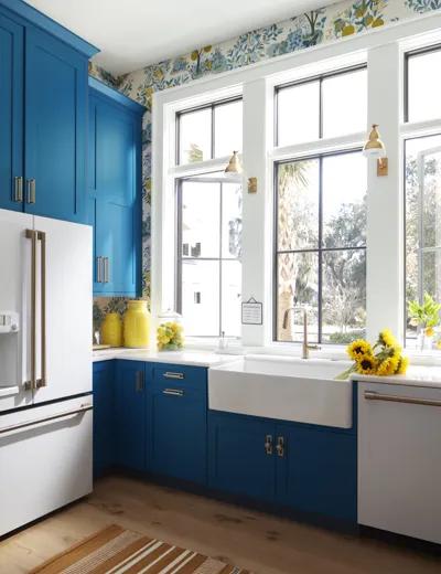 Una cocina con gabinetes pintados en azul, techo y marcos blancos, y papel pintado floreado sobre una pared con ventanas.