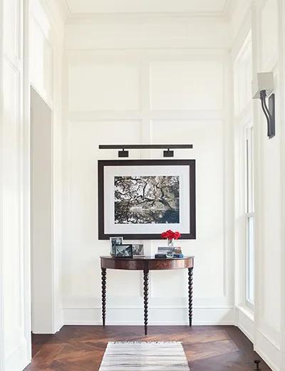 Un pasillo pintado de blanco con paredes con revestimientos y tablas, un techo blanco, marco blanco y arte enmarcado por encima de una mesa.