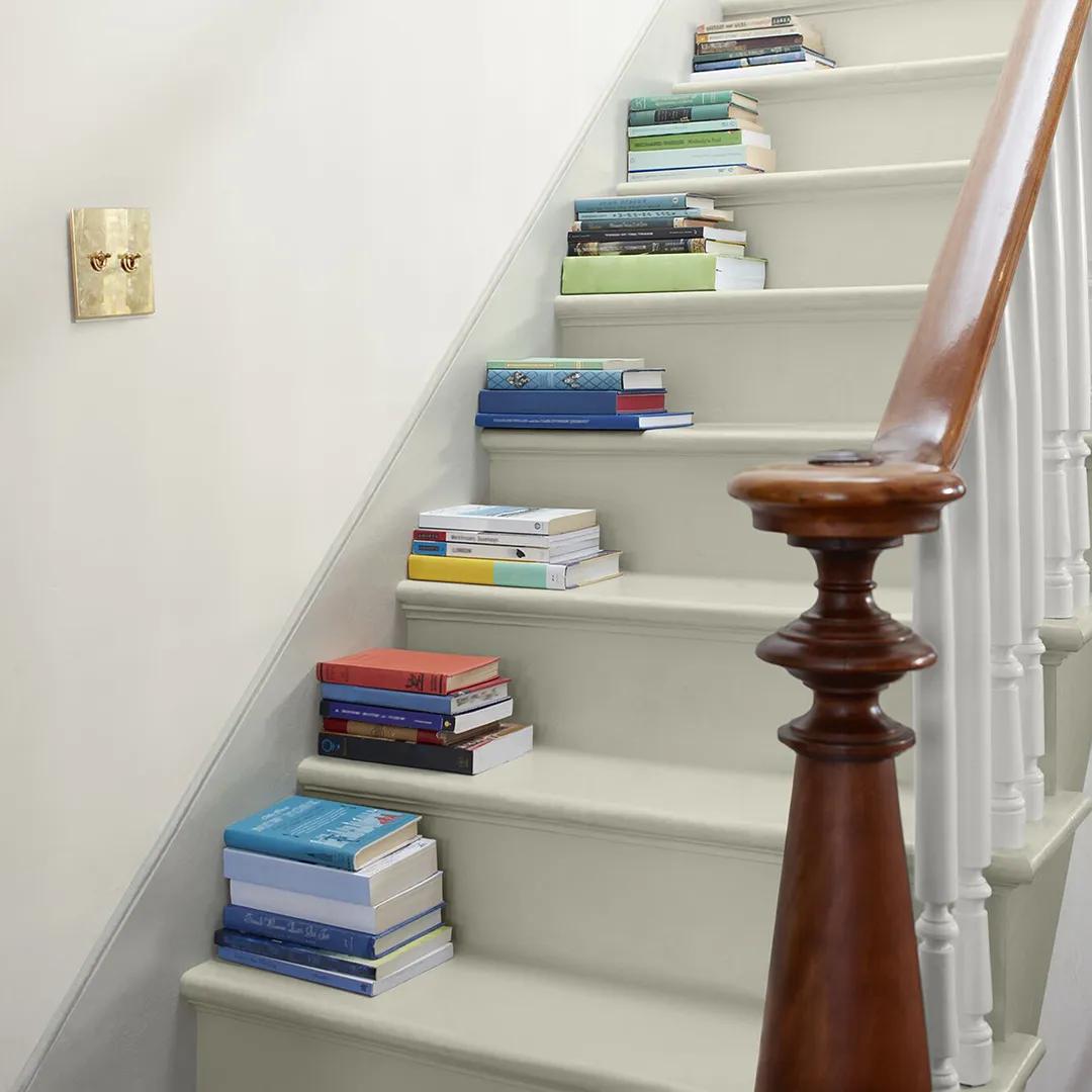 Una escalera pintada de blanco y blanco impuro y pared con un pasamanos de madera y un poste central, y libros apilados en cada escalón.