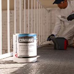 Miniatura: Una lata de 1 galón de Command® Uretano acrílico al agua de Benjamin Moore junto a un contratista de pintura pintando una barandilla blanca.