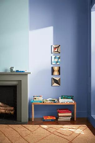La esquina de una sala de estar con una pared pintada en un azul claro y nítido, una chimenea, una alfombra naranja y un banco lleno de libros debajo de cuatro pequeñas obras de arte.