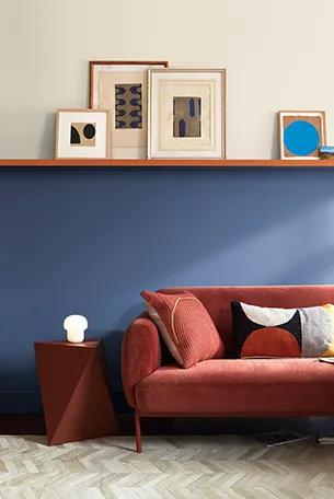 Una sala de estar con una pared dividida por una repisa pintada en un azul suntuoso con toques de violeta en la mitad inferior y un tono blanco impuro en la pared superior, una repisa que sostiene varios marcos y un sofá de terciopelo naranja con cojines gráficos.