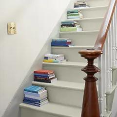 Miniatura: Una escalera pintada de blanco y blanco impuro y pared con un pasamanos de madera y un poste central, y libros apilados en cada escalón.