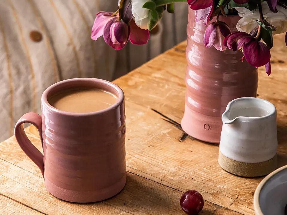 Un jarrón con flores rosas y una taza de cerámica en marrón tenue sobre una mesa de madera.