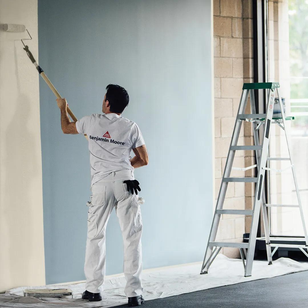 Un contratista de pintura profesional aplica pintura blanca con un rodillo sobre una pared interior azul.