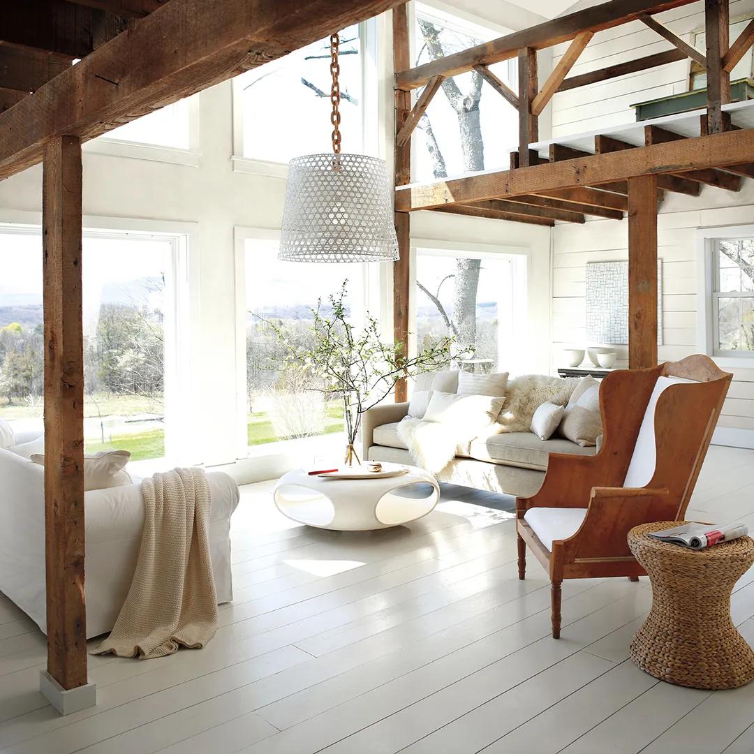 Una sala de estar luminosa pintada de blanco con ventanas altas, vigas de madera natural, un área de loft y decoración informal.